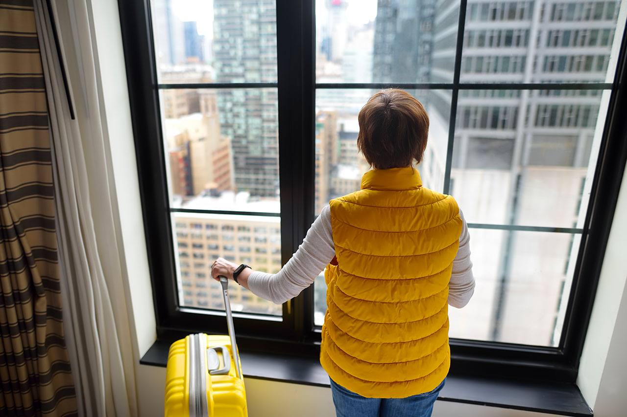 Voyageuse en doudoune jaune avec sa valise, devant la fenêtre d'une chambre d'hôtel donnant sur des gratte-ciel